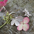 Silberschmuck mit Blattstrukturen neben einer rosa Blüte